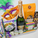 Mardi Gras-themed gift basket with champagne, snacks, and a mask on a white background