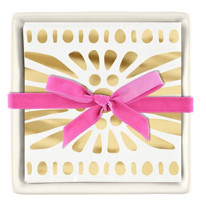 Decorative paper cocktail napkins featuring a metallic gold sunburst pattern are displayed in a square white ceramic tray and tied with a vibrant pink velvet ribbon bow