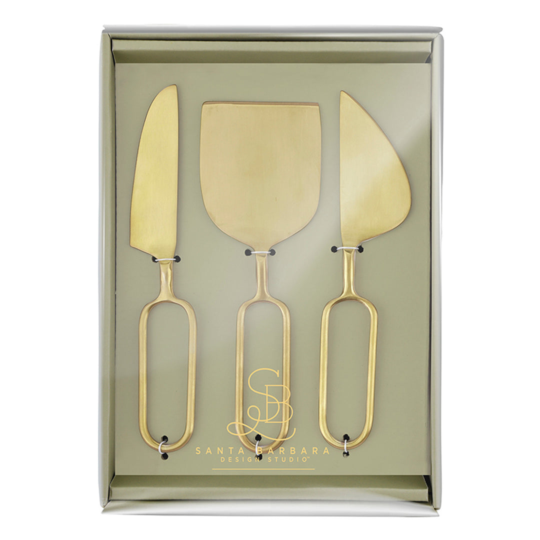 Brass-finished stainless steel cheese tool set by Santa Barbara Design Studio featuring three distinct serving knives with minimalist elongated oval loop handles displayed inside a sage-green gift box with a gold-foil logo