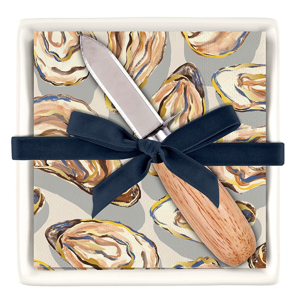 Painterly illustration by Caspari of an oyster-patterned cocktail napkin and wooden-handled knife gift set featuring a navy blue ribbon bow and gold-flecked shell motifs inside a white ceramic dish