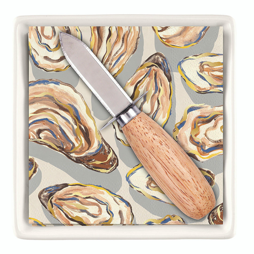 Digital product photograph by Michel Design Works of a stainless steel oyster knife with a light wood handle resting on paper napkins featuring painterly blue and yellow oyster shell illustrations inside a white square ceramic tray
