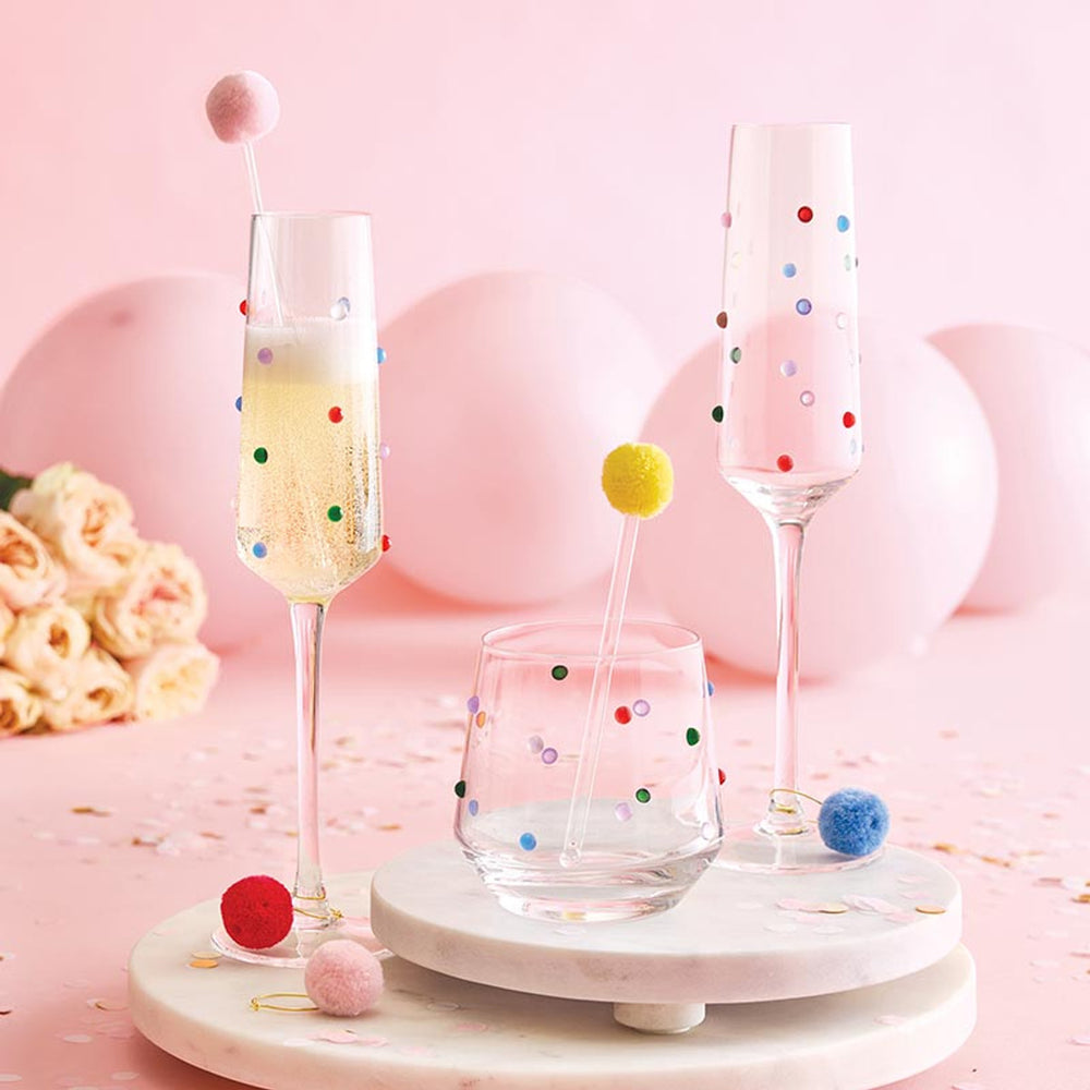 Studio photograph of champagne flutes and a lowball glass adorned with vibrant multi-colored dots set on white marble platters against a festive backdrop of pink balloons, soft roses, and whimsical pom-pom drink stirrers