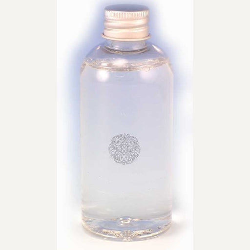 Photograph of a transparent bottle of clear liquid positioned in a minimalist white studio setting featuring a ribbed metallic silver cap and an intricate circular filigree emblem centered on the body