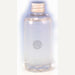 Photograph of a transparent bottle of clear liquid positioned in a minimalist white studio setting featuring a ribbed metallic silver cap and an intricate circular filigree emblem centered on the body