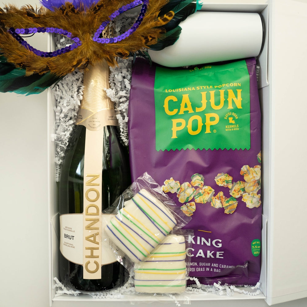 Photograph of a Mardi Gras celebration gift box containing a bottle of Chandon Brut, a purple bag of Cajun Pop King Cake popcorn, and a vibrant gold feathered mask with purple sequins alongside striped white cookies