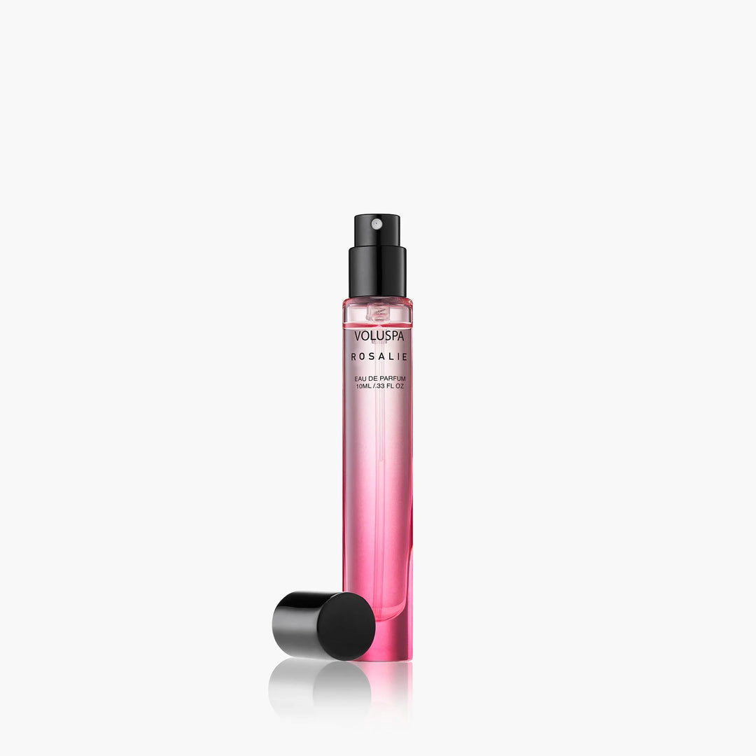 Product photograph of Voluspa Rosalie Eau de Parfum travel spray on a white studio background featuring a slim pink glass bottle with a black spray nozzle and a detached cap lying beside its soft reflection
