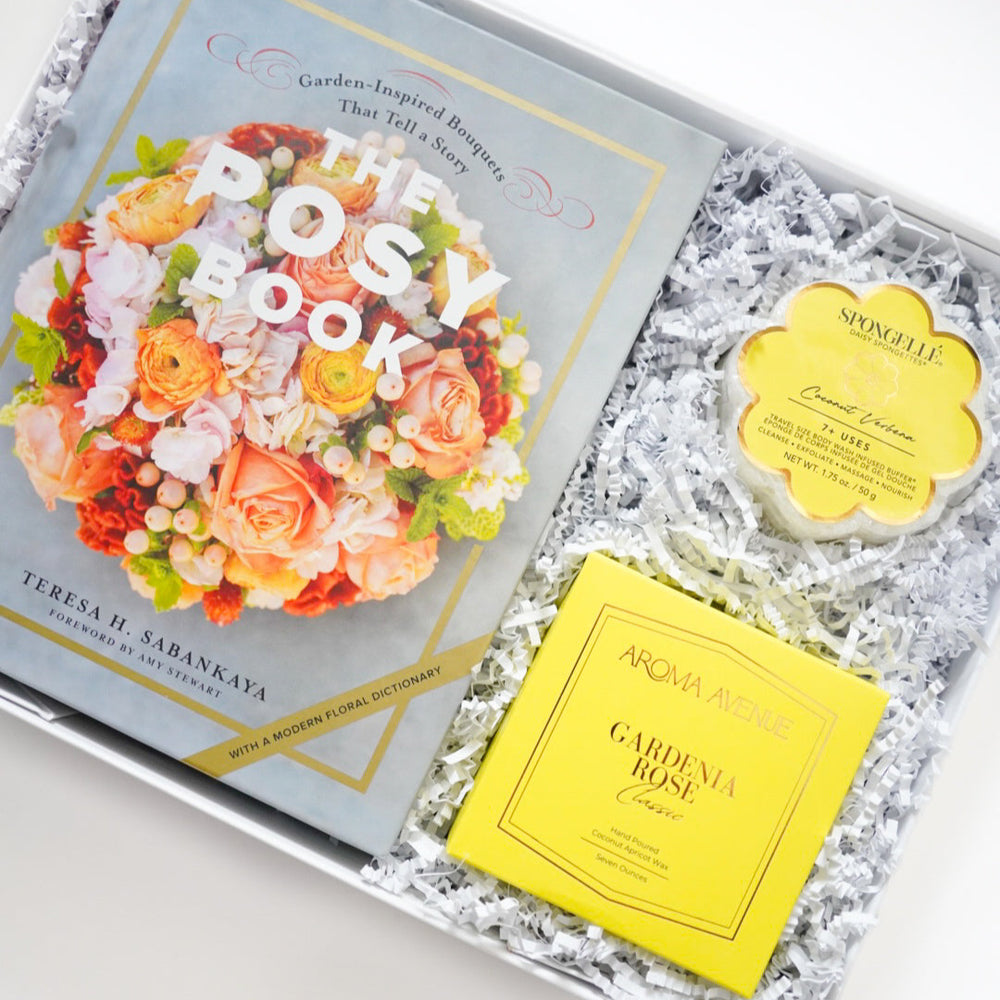 Photograph of Teresa H. Sabankaya's The Posy Book with a vibrant orange floral cover in a curated gift box alongside a yellow Aroma Avenue candle and flower-shaped Spongellé buffer resting on white crinkle paper