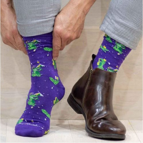 Photograph of Gold Standard socks showing a person in grey trousers wearing purple socks decorated with green alligators playing golden saxophones and a brown leather boot with a side zipper on a light wooden background