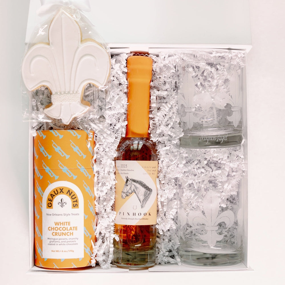 Photograph of Mignon Faget etched fleur-de-lis glassware and a Pinhook bourbon bottle with an orange wax seal arranged in a New Orleans gift box featuring a blue trumpet-patterned canister and white chocolate crunch treats