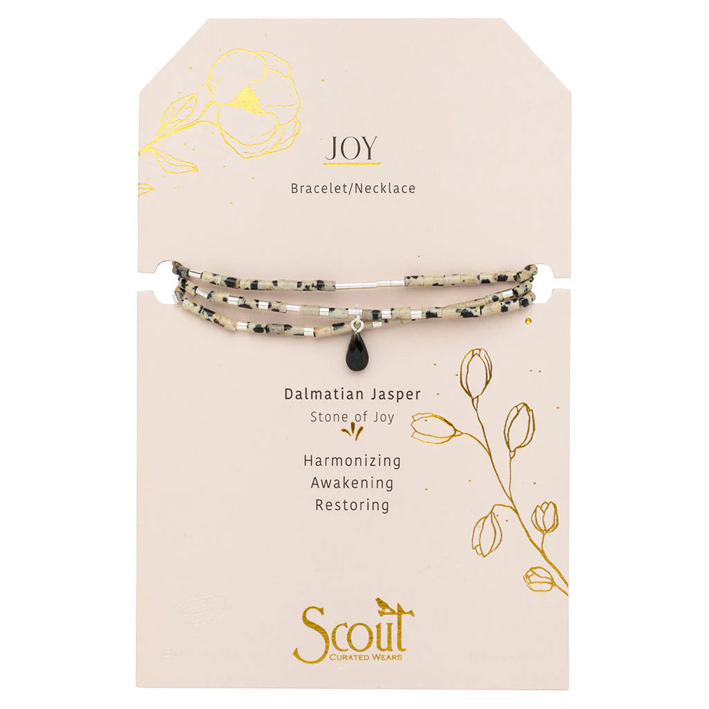 Jewelry by Scout Curated Wears features a Dalmatian Jasper bracelet necklace with spotted beige beads and a black teardrop charm on a card decorated with gold flowers and the word JOY