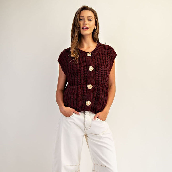 Woman wearing a burgundy chunky knit vest with large gold buttons and white wide-leg jeans