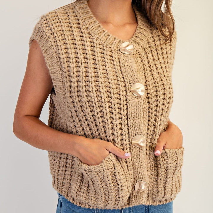 Close-up of a tan chunky knit vest with sculptural gold buttons, worn with hands in the pockets