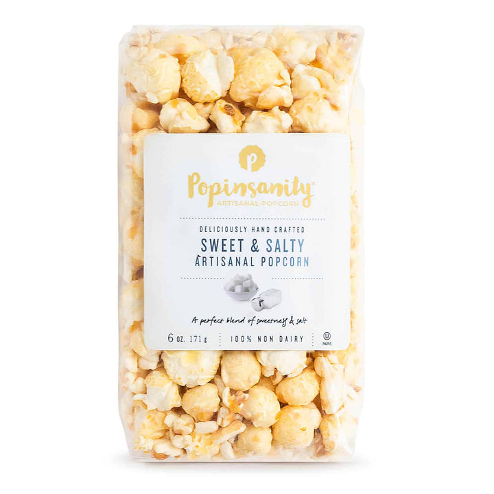 Commercial product photograph by Popinsanity of a clear bag of Sweet &amp; Salty Artisanal Popcorn on a white studio background featuring large golden kernels, a signature gold P logo, and blue serif typography