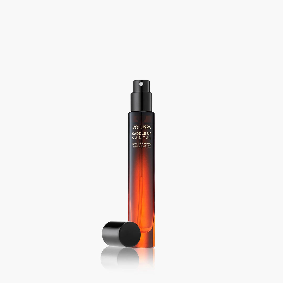 Product photograph by Voluspa of a slender Saddle Up Santal Eau de Parfum travel bottle on a white background featuring warm amber gradient glass, a black spray nozzle, and a detached cap resting beside its soft reflection