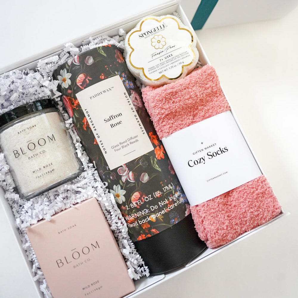 Product photograph by Giften Market of a curated spa gift box featuring Bloom Bath Co. wild rose bath soak, a floral Paddywax Saffron Rose diffuser, a flower-shaped Spongellé buffer, and plush pink fuzzy socks nestled in white crinkle paper