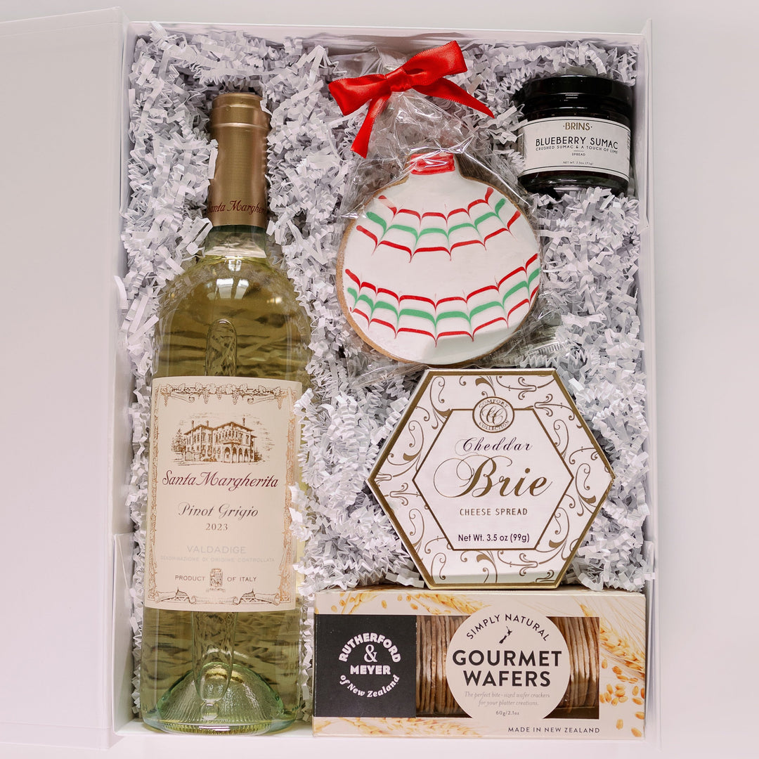 Photograph of a luxury holiday gift box featuring a bottle of Santa Margherita Pinot Grigio with its iconic villa label, an intricately iced red and green ornament cookie with a festive bow, and a hexagonal box of cheddar brie cheese spread nestled in white crinkle paper