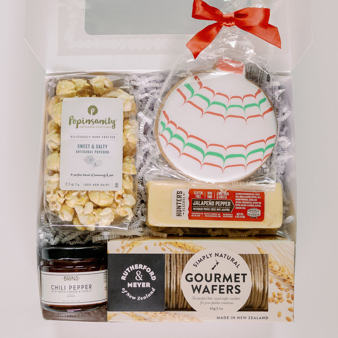 Photograph of a curated holiday food gift box featuring an ornament-shaped sugar cookie with red and green zig-zag icing, a vibrant red satin bow, and gourmet items like Popinsanity popcorn and Rutherford and Meyer wafers from New Zealand