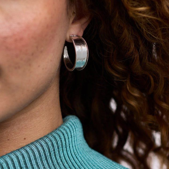 Digital photograph of a wide silver hoop earring with beaded edges worn by a woman with freckled skin and a vibrant turquoise ribbed turtleneck sweater