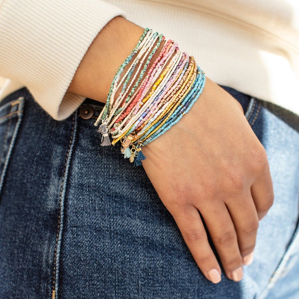 Photograph of a bohemian-style multi-strand bracelet stack worn on a wrist featuring tiny turquoise and coral seed beads with small hanging tassels against blue denim jeans