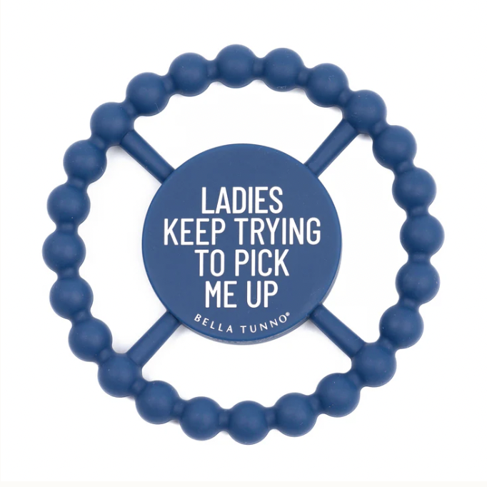 Silicone teether by Bella Tunno featuring a navy blue circular baby toy on a white background with a central disc displaying the text Ladies Keep Trying To Pick Me Up and a textured beaded outer ring connected by four spokes
