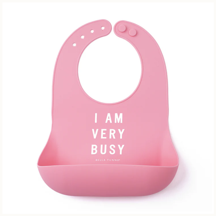 Silicone baby bib by Bella Tunno featuring a pink surface with white text reading I AM VERY BUSY and featuring an adjustable button neck closure alongside a wide food-catcher pocket against a white background