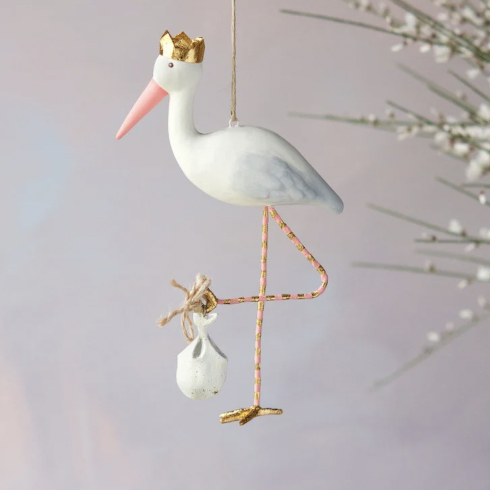 Hand-painted hanging ornament of a white stork wearing a gold crown and pink beak while carrying a white bundle on its pink and gold striped legs against a neutral backdrop with white flowers