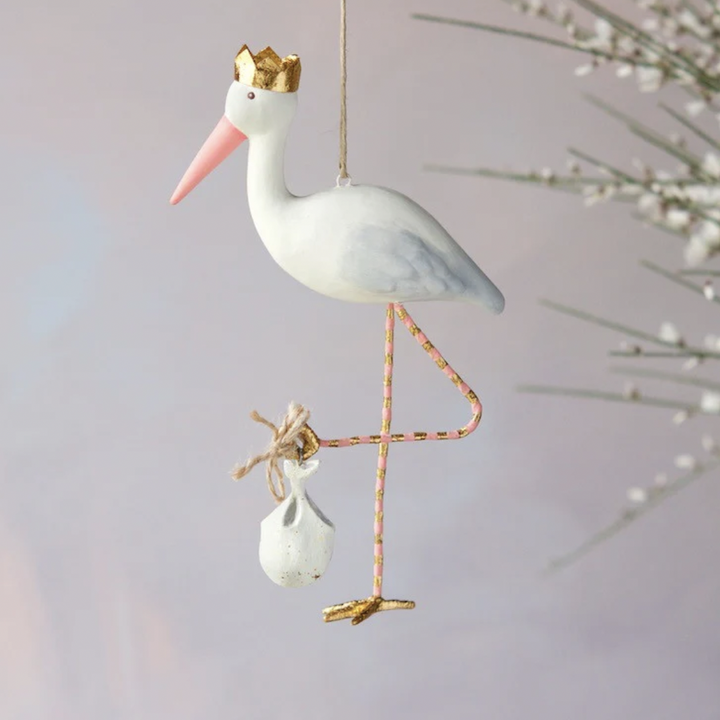 Hand-painted hanging ornament of a white stork wearing a gold crown and pink beak while carrying a white bundle on its pink and gold striped legs against a neutral backdrop with white flowers