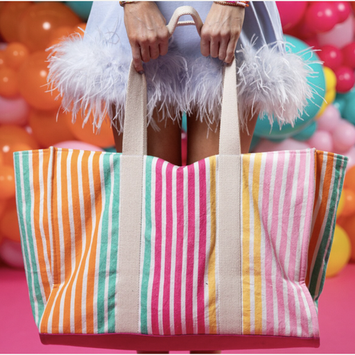 Digital photograph of a large canvas tote bag with vibrant orange, teal, and pink vertical stripes held by a person wearing a light blue dress with fluffy ostrich feather trim against a festive backdrop of multicolored balloons
