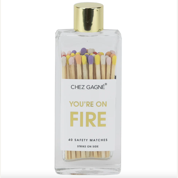 Product photograph by Chez Gagné of a clear glass bottle containing 40 safety matches with multicolored heads and a gold cap, featuring a white label with the text YOU'RE ON FIRE in yellow font against a plain white background