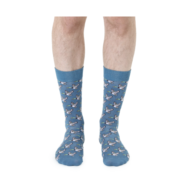 Photograph of blue patterned crew socks worn by a person against a white background featuring a repeating motif of flying mallard ducks with vibrant green heads and white-tipped wings on a dusty blue fabric