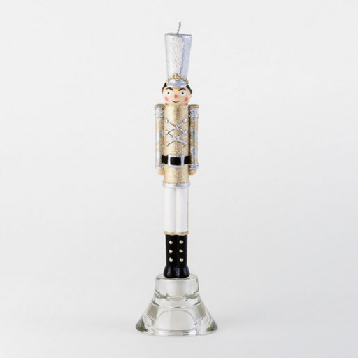Hand-painted wax nutcracker soldier candle standing in a glass holder against a white background featuring a shimmering gold glitter uniform, a tall silver shako hat, and black boots with gold accents