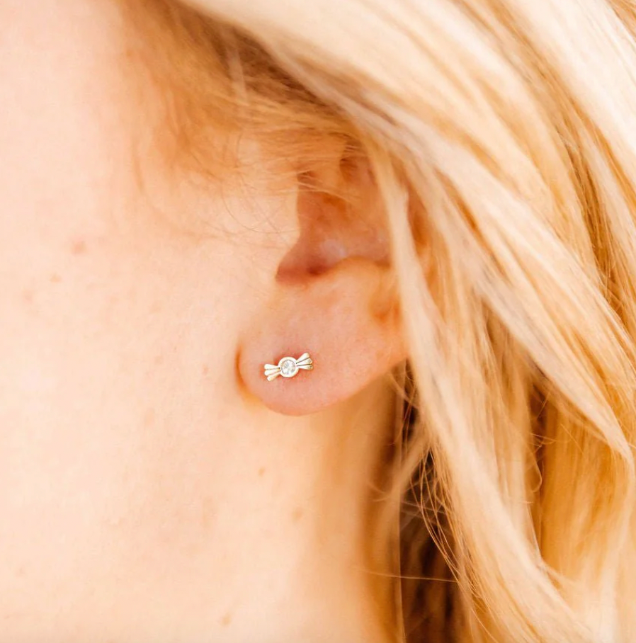 Digital photograph of a small gold candy-shaped earring with a central sparkling diamond on a woman's earlobe, accented by wispy blonde hair and soft skin tones