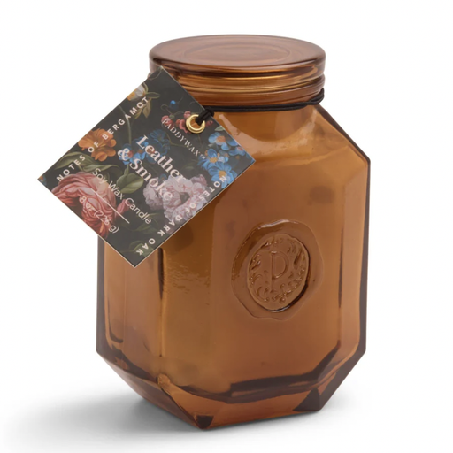 Studio photograph by Paddywax of a Leather and Smoke soy wax candle in an amber-tinted hexagonal glass jar featuring an embossed P seal and a vibrant floral hangtag against a plain white background