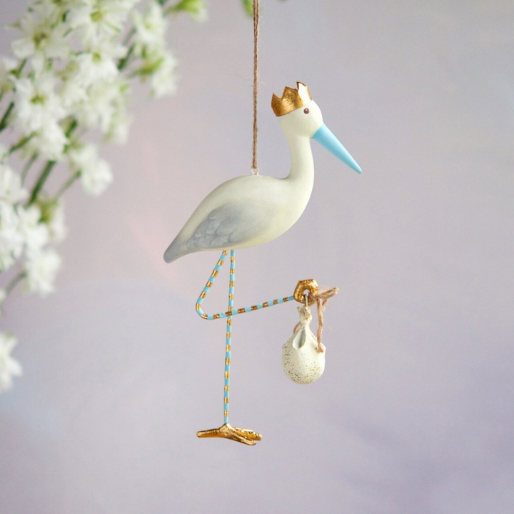 Handcrafted painted stork ornament hanging against a soft white floral backdrop, featuring a gold-crowned head, a light blue beak, and blue and gold striped legs carrying an iconic baby bundle