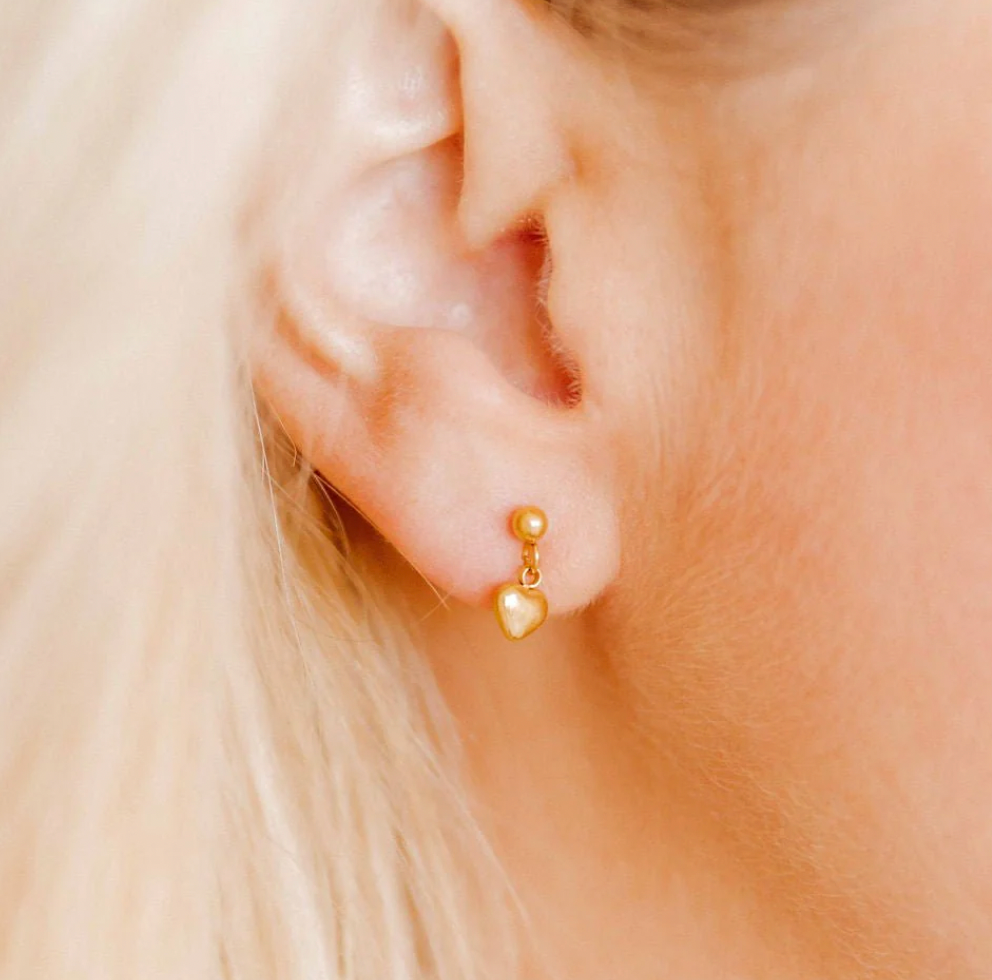 Photograph of a dainty gold heart drop earring worn on a human earlobe featuring a polished metallic finish and a small round stud nestled against soft blonde hair