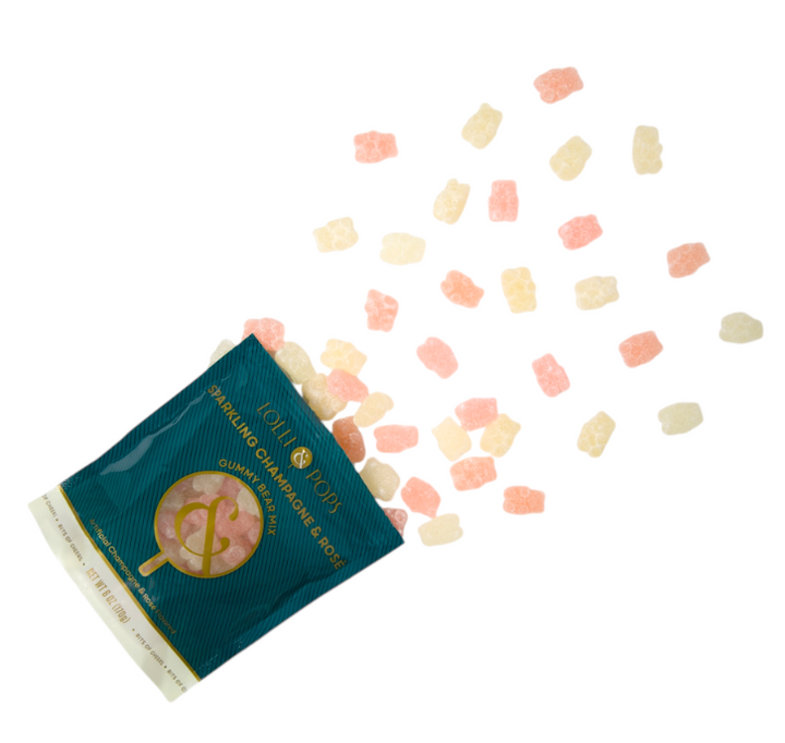 Studio photograph by Lolli &amp; Pops of a teal bag of Sparkling Champagne and Rosé gummy bears spilling across a plain white surface with translucent pink and cream candies and gold foil typography