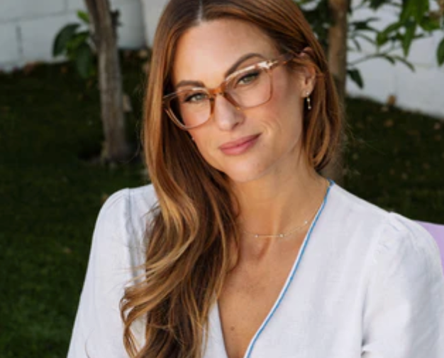 Woman with long auburn hair wearing translucent tan glasses and a white top outdoors