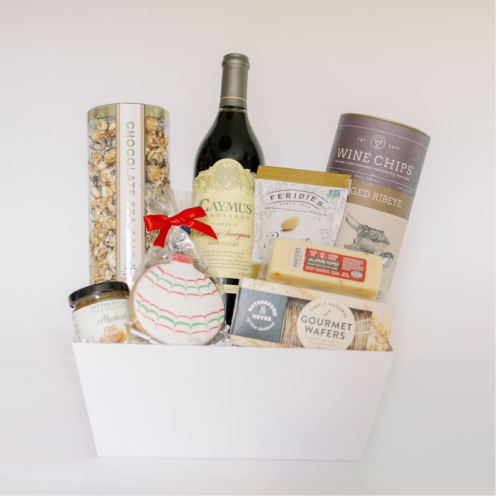 Photograph of a gourmet gift basket on a white background featuring a bottle of Caymus Napa Valley Cabernet Sauvignon, a canister of Wine Chips, and a round frosted cookie with a bright red ribbon bow