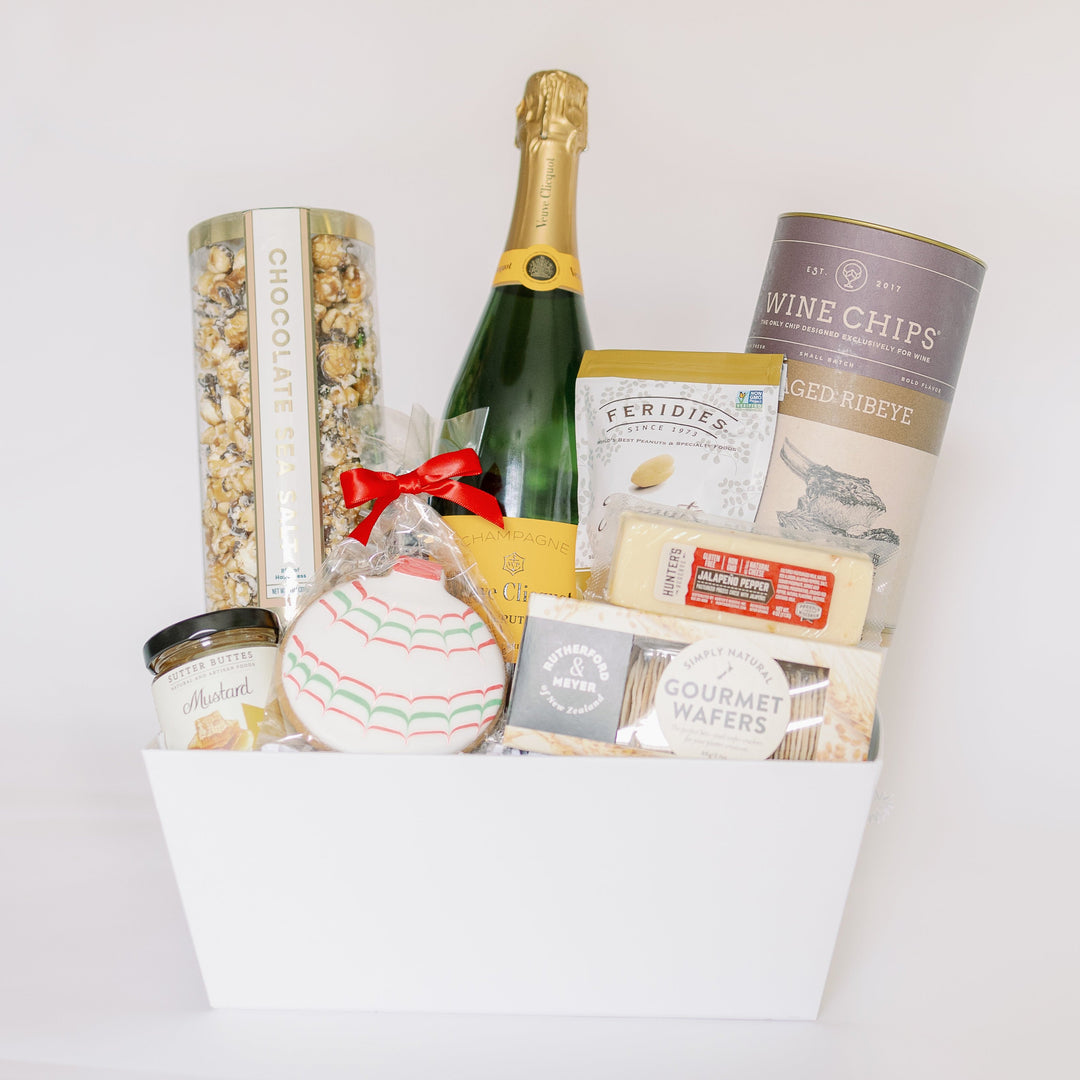 Professional studio photograph of a luxury holiday gift basket in a white box featuring a signature yellow-labeled Veuve Clicquot champagne bottle, a festive hand-iced ornament cookie with a red ribbon, and gourmet snacks like chocolate sea salt popcorn and aged ribeye wine chips