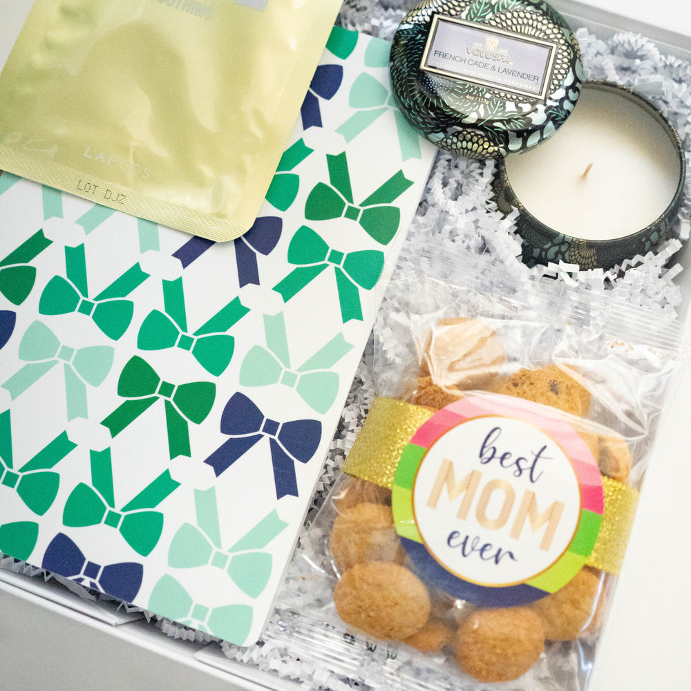 Photograph of a Mother's Day gift box featuring a Voluspa French Cade & Lavender candle and Lapcos face mask alongside cookies with a Best Mom Ever rainbow label and teal bow-patterned stationery over white crinkle paper