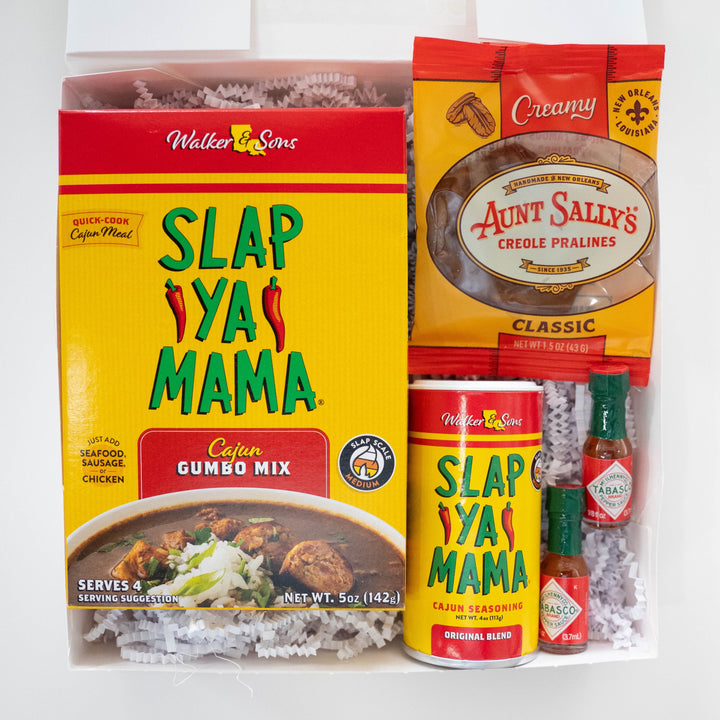 Product photograph of a New Orleans culinary gift box featuring iconic Slap Ya Mama gumbo mix and seasoning, Aunt Sally’s Creole Pralines, and two tiny miniature Tabasco bottles nestled in white crinkle-cut paper filler