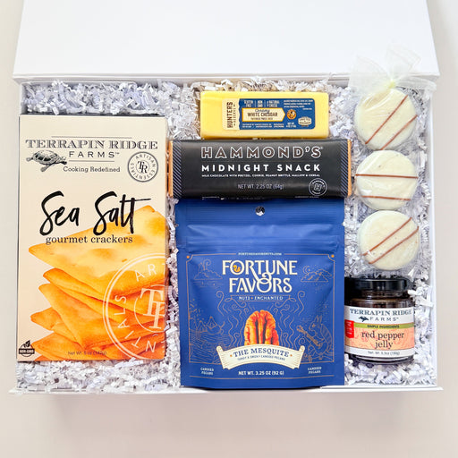 An open gift box filled with assorted snacks and treats, including Terrapin Ridge Farms Sea Salt chips, Diamond's Midnight Snack chocolate, Fortune Favors blue bag, and a small jar, arranged in white crinkle paper on a light background.