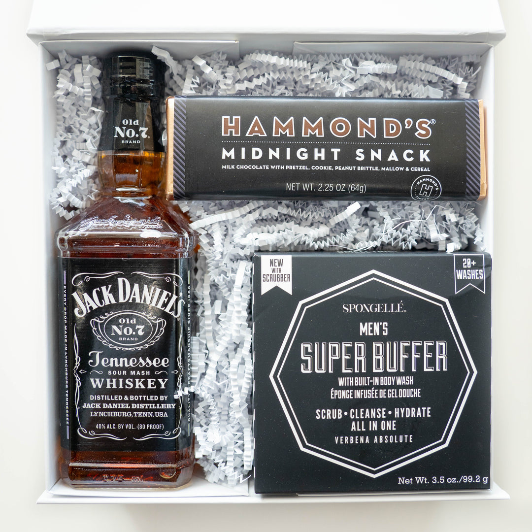 A studio photograph of a curated men's gift box featuring a glass bottle of amber Jack Daniel's Tennessee Whiskey, a Hammond's Midnight Snack chocolate bar with gold foil ends, and a black Spongellé Super Buffer box resting on white crinkle paper filler