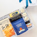 An open white gift box tied with a blue ribbon, filled with assorted gourmet snacks and treats on a light beige background. Visible items include a box of Heaven's Ridge Sea Salt, a blue bag of Fortune Fancies, a dark chocolate bar, and several macarons.