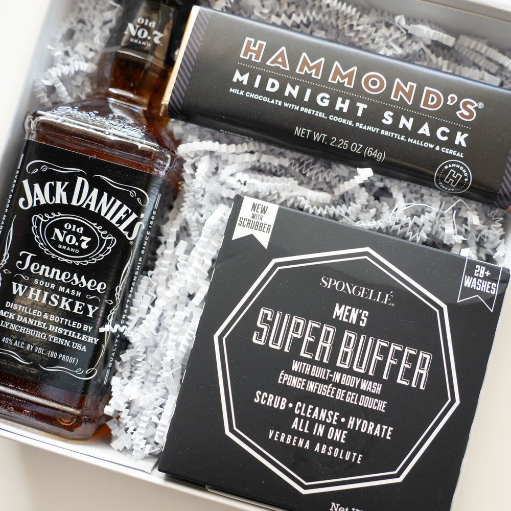 Digital photograph of a curated men's gift box featuring iconic Jack Daniel’s Tennessee Whiskey, a Hammond’s Midnight Snack chocolate bar, and a Spongellé Men’s Super Buffer resting on textured white zig-zag crinkle paper filler