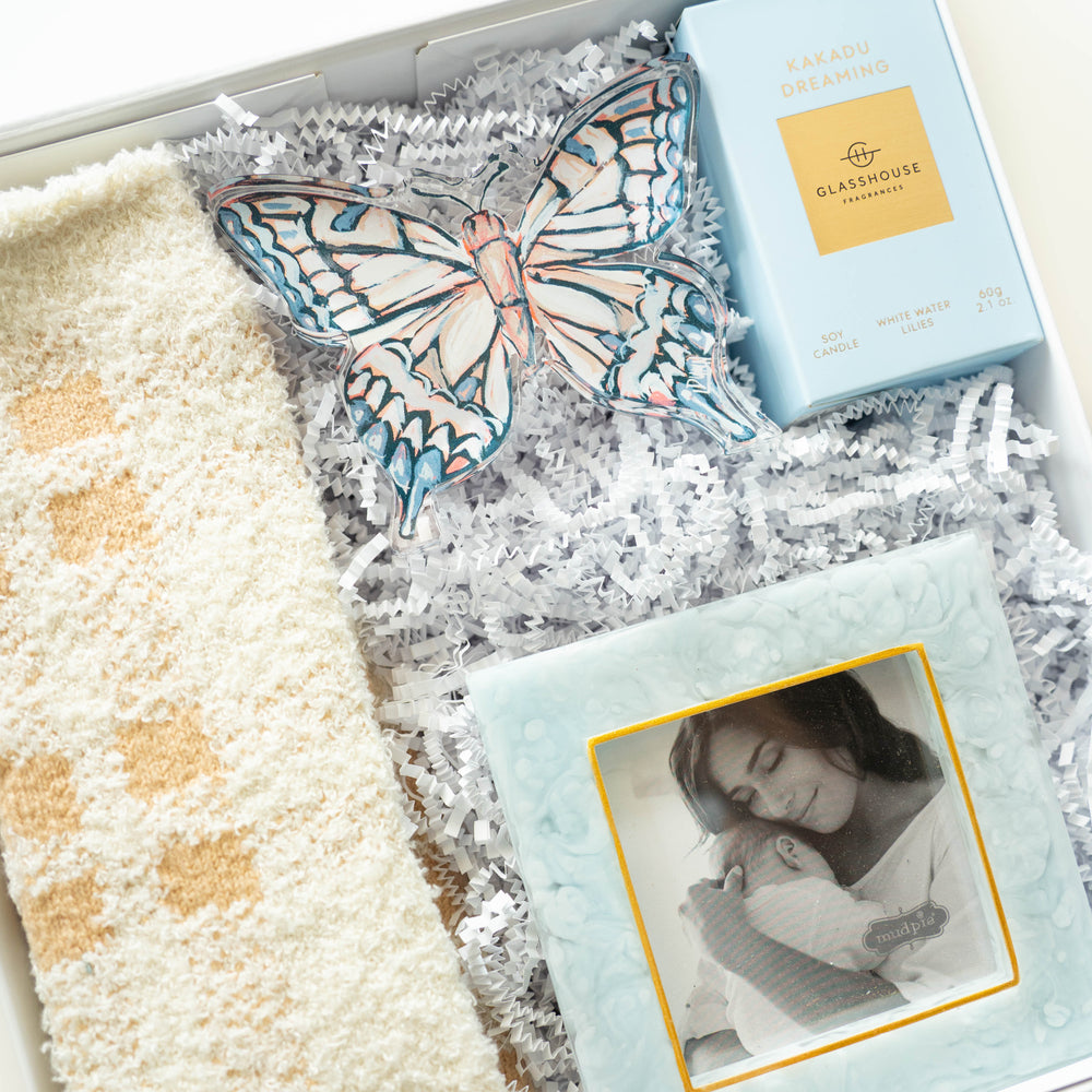 Photograph of a curated gift set featuring a Glasshouse Fragrances Kakadu Dreaming candle and a Mud Pie photo frame containing a black and white portrait of a mother and baby alongside an intricate blue and orange patterned acrylic butterfly and a plush cream blanket nestled in white crinkle paper