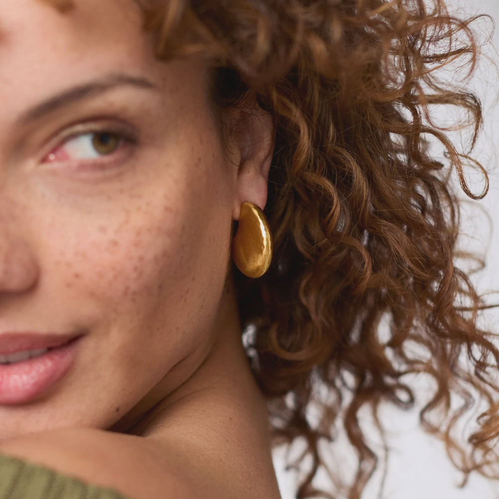 Photograph of a woman with freckled skin and voluminous brown curls wearing a polished teardrop-shaped gold earring in a close-up studio shot