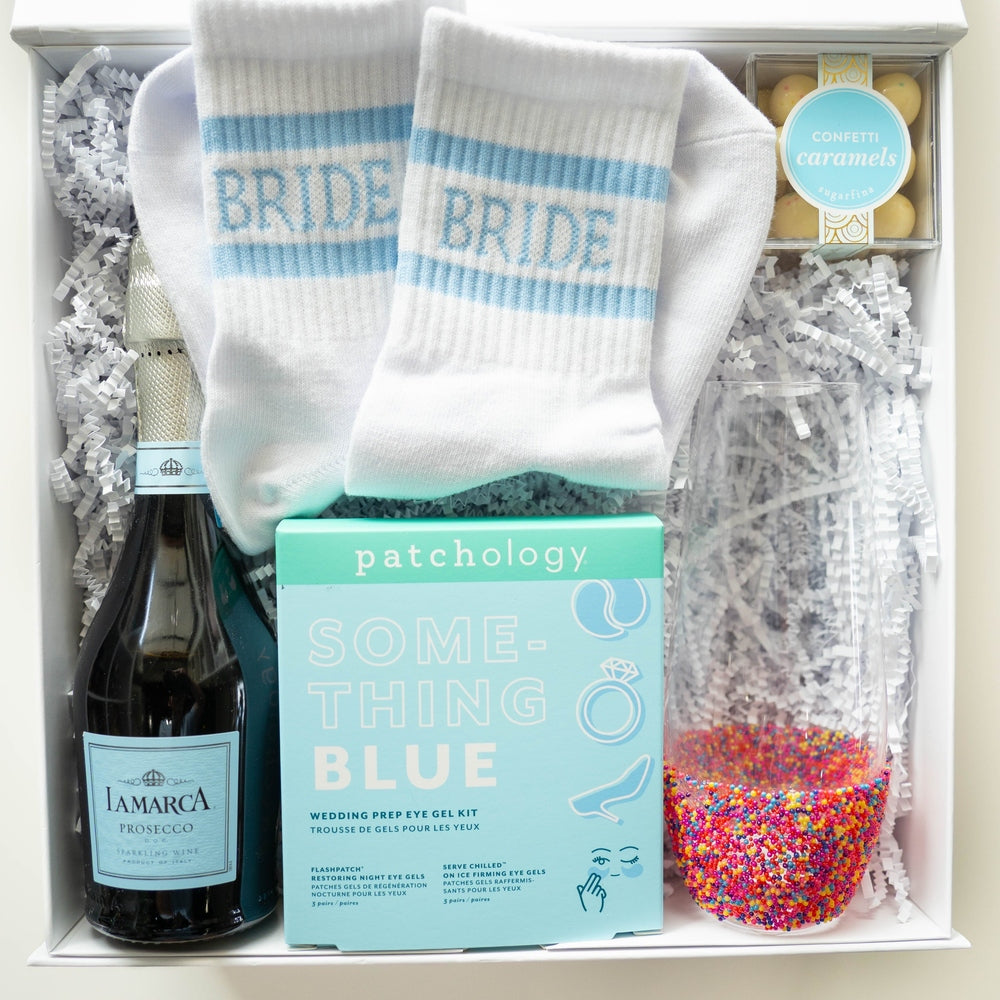Photograph of a curated bridal gift box featuring a bottle of La Marca Prosecco, a light blue Patchology eye gel kit, Sugarfina confetti caramels, white socks with blue Bride lettering, and a stemless glass filled with colorful rainbow sprinkles