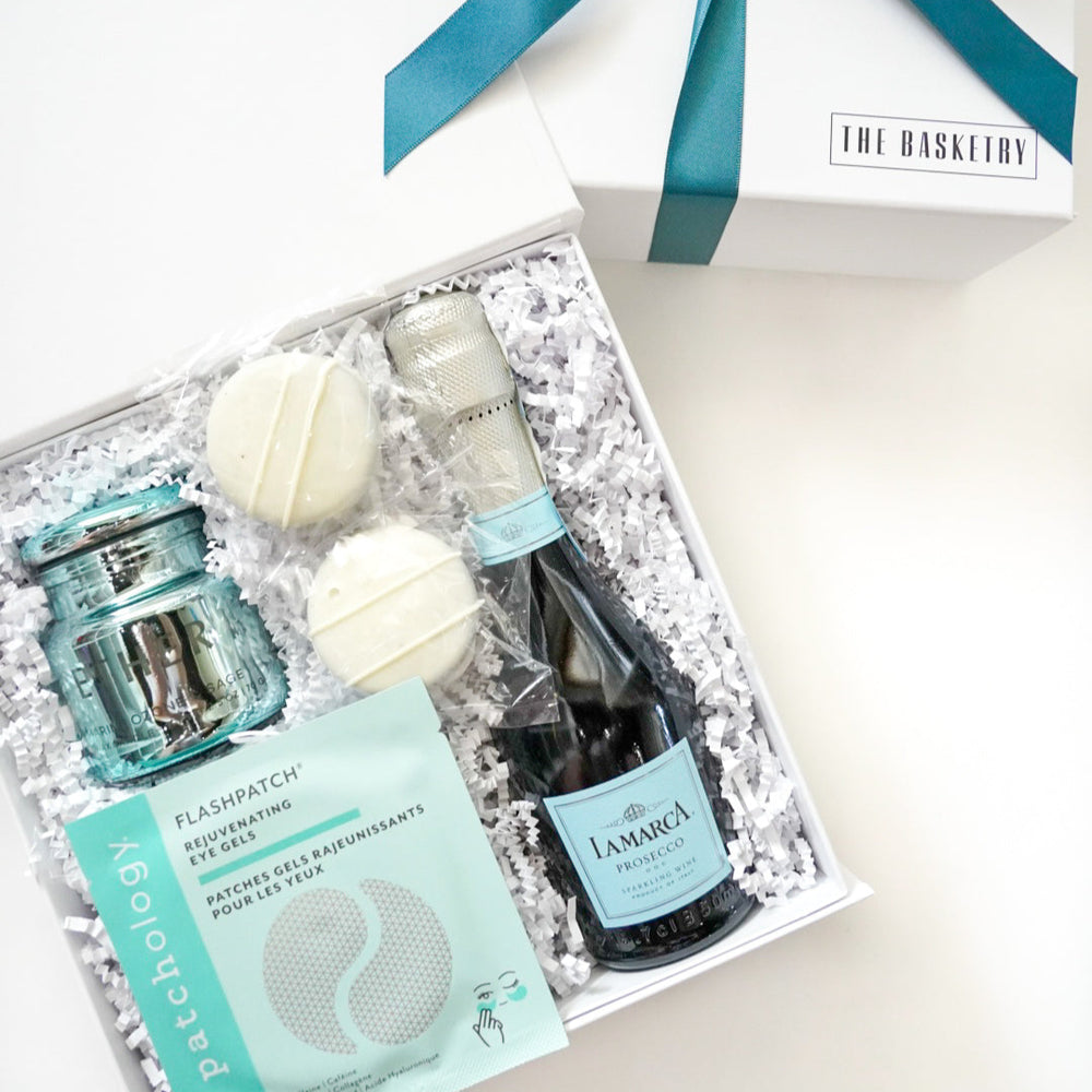 Photograph by The Basketry of a white gift box featuring a mini bottle of La Marca Prosecco, light blue Patchology eye gels, and a turquoise glass jar nestled in white shredded paper and accented with a teal ribbon