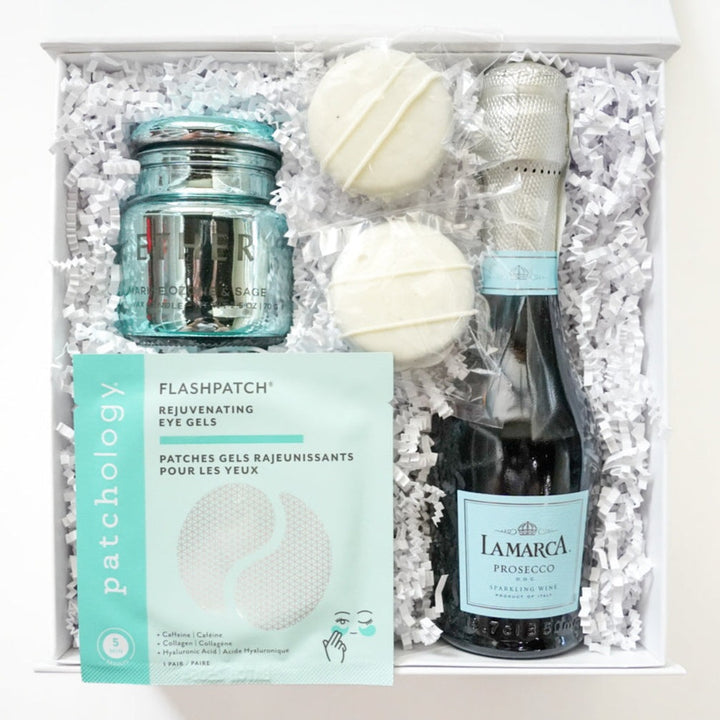 Photograph of a curated self-care gift box featuring a mini bottle of La Marca Prosecco with its iconic light blue label, a teal glass Ether candle, and two white chocolate-covered cookies nestled in white shredded paper